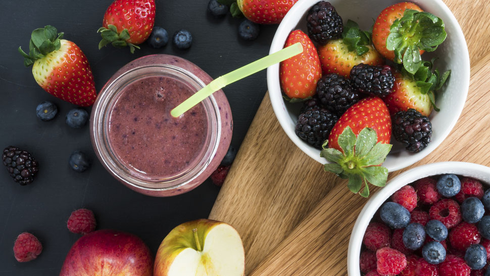 A berry smoothie in a jar surrounded by half an apple and a bowl of berries.