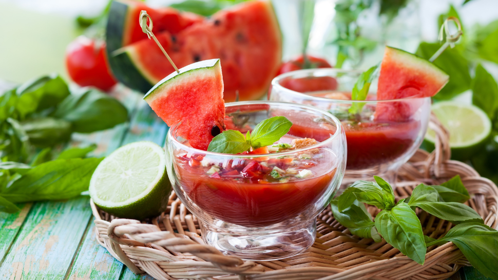 Watermelon gazpacho served in glass bowls with basil garnish, lime halves, and fresh watermelon slices.