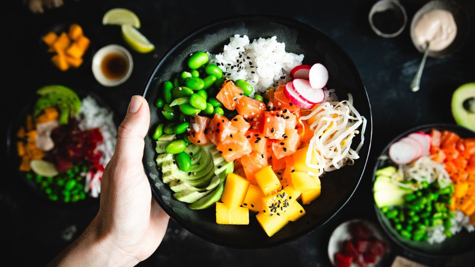 A hand holds a vibrant poke bowl with salmon, mango, avocado, edamame, radish, bean sprouts, and rice, garnished with black sesame seeds.