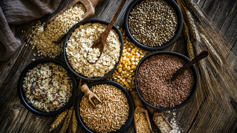 An assortment of grains in black bowls on a wooden surface, including oats, corn, and flaxseeds. Rustic spoons add a natural, wholesome feel.