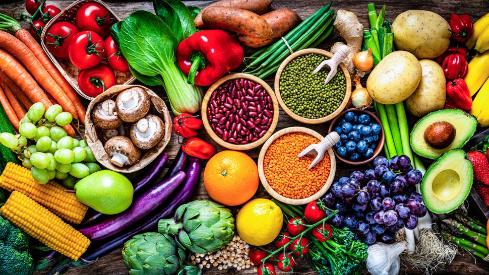 A vibrant array of fruits, vegetables, and legumes on a table. The spread includes tomatoes, grapes, carrots, beans, and avocados, conveying freshness.