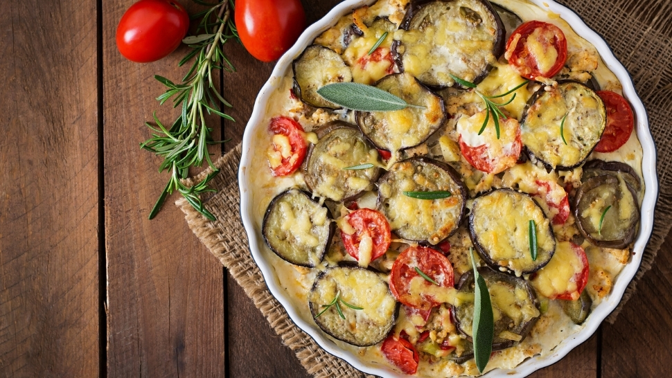 vegetarian moussaka in a white baking dish on a wood table with tomatoes and herbs
