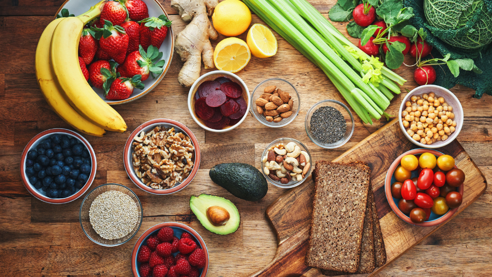 Assorted fruits, vegetables, nuts, grains, and bread arranged on a wooden table, including bananas, berries, avocado, chickpeas, and tomatoes.