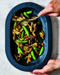 Hands holding a platter of stir-fried oyster mushrooms with snap peas and peppers.