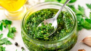 Fresh chimichurri in a glass jar with herbs, garlic, and oil on a light surface.