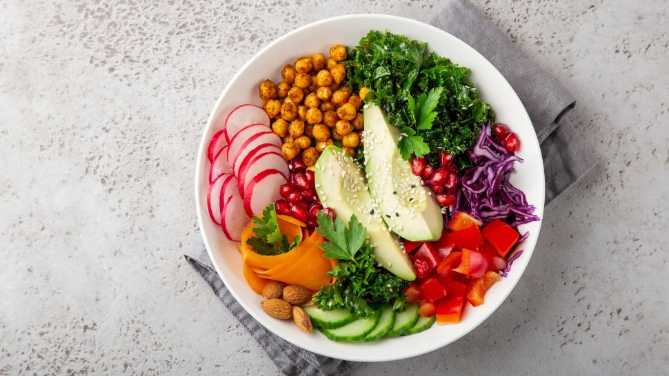 Colorful grain bowl with avocado, chickpeas, kale, radishes, cabbage, and fresh vegetables arranged in sections.