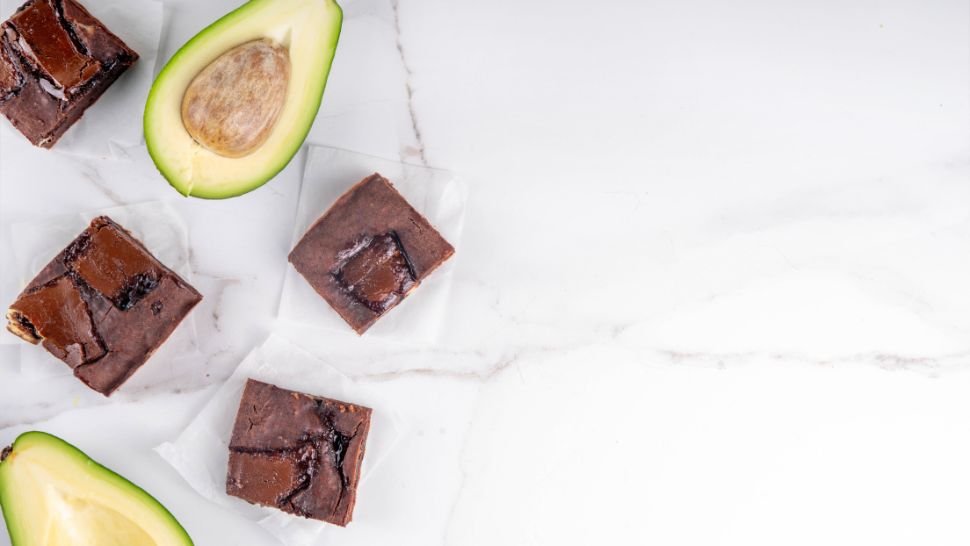 Slices of chocolate brownies and halved avocados on a marble surface.