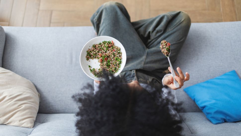 Person sitting on a couch holding a bowl of tabbouleh salad and a spoon, with crossed legs and a blue cushion nearby.