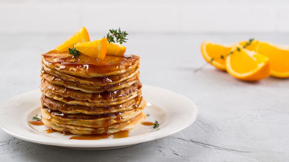 Fluffy pancakes with orange wedges and maple syrup, styled with a sprig of thyme.