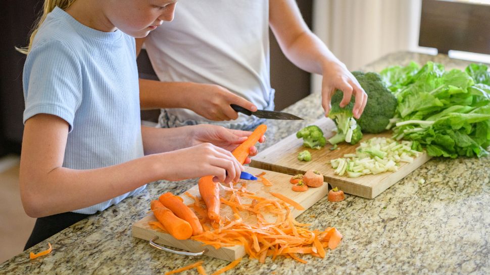 Kids peel and chop garden vegetables on a sunlit kitchen counter.