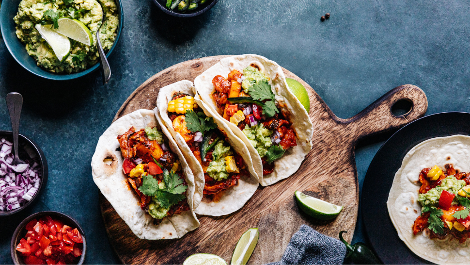 Three tacos with grilled vegetables, corn, and guacamole on a wooden board, surrounded by lime wedges and toppings.
