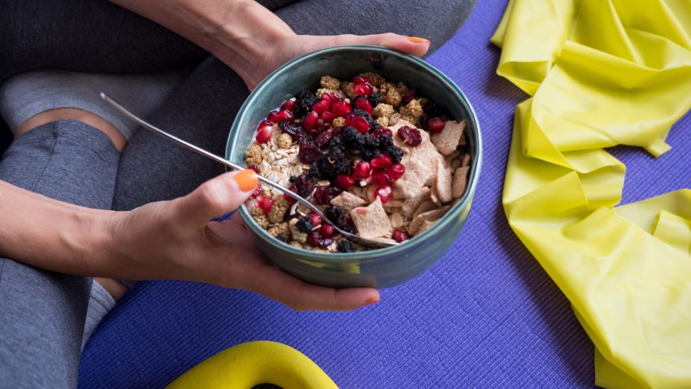 Person holding a bowl of oats with berries and pomegranate on a yoga mat.
