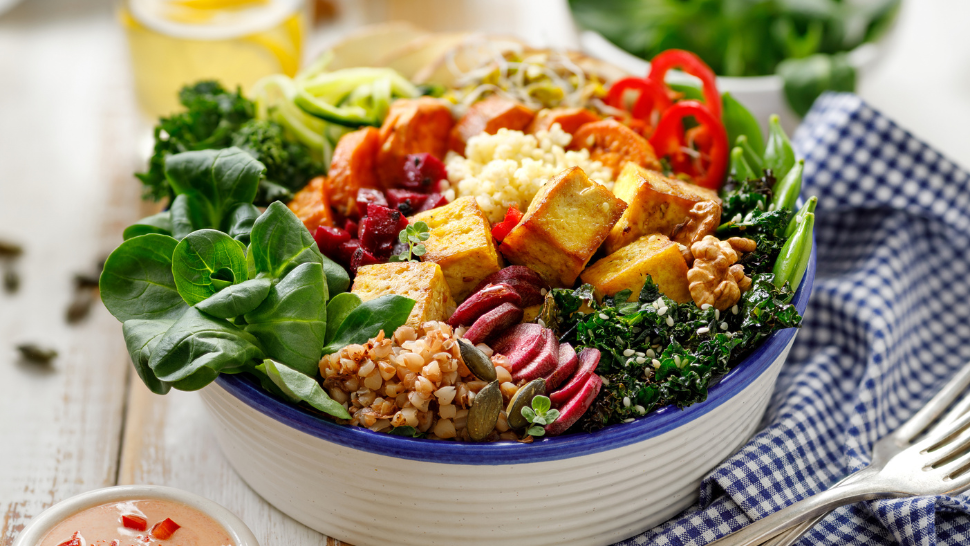 Bowl of plant-based meal with tofu, leafy greens, grains, roasted vegetables, and seeds arranged in colorful sections.