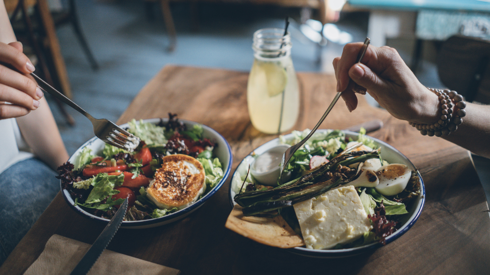 Two people sharing colorful salads with grilled vegetables and cheese at a wooden table, practicing mindful eating together.