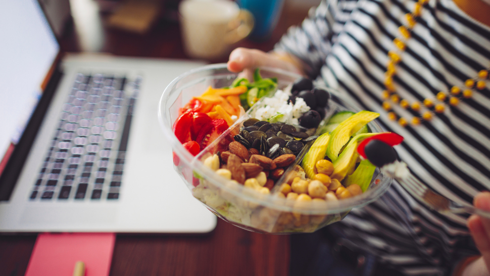 Person holding a colorful grain and vegetable bowl while working at a laptop, highlighting balanced eating during a busy workday.