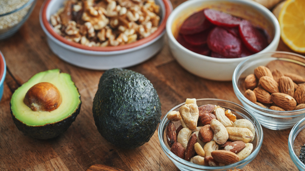Avocados, nuts, and sliced beets arranged on a wooden table with bowls of healthy snack ingredients.