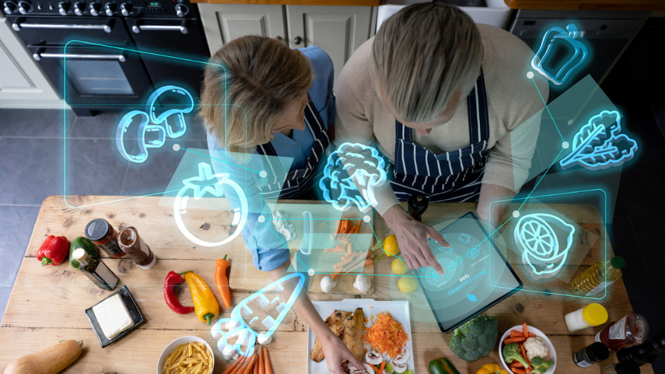 Two people cooking in a kitchen using a tablet app, with digital food icons overlaying fresh ingredients on the counter.