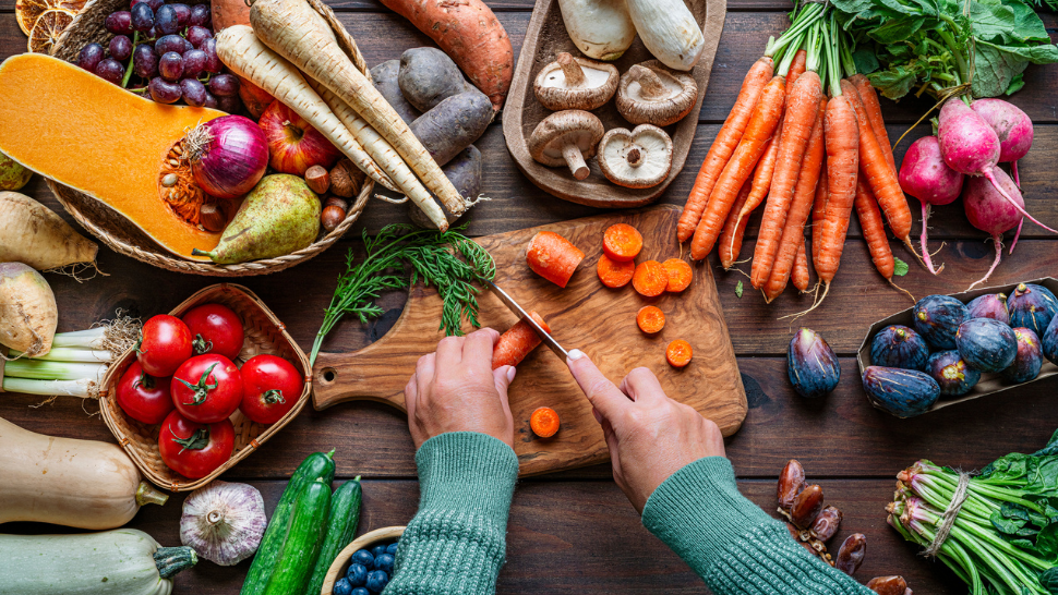 Hands chopping carrots amid a spread of seasonal root vegetables, fruits, and herbs on a rustic table.