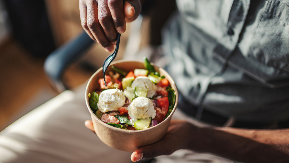 Person holding a bowl of fresh salad with tomatoes, cucumbers, greens, and scoops of soft cheese.