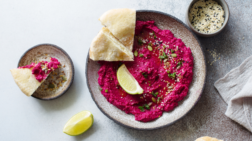 Bowl of vibrant beet hummus topped with seeds and herbs, served with pita triangles and lime wedges.