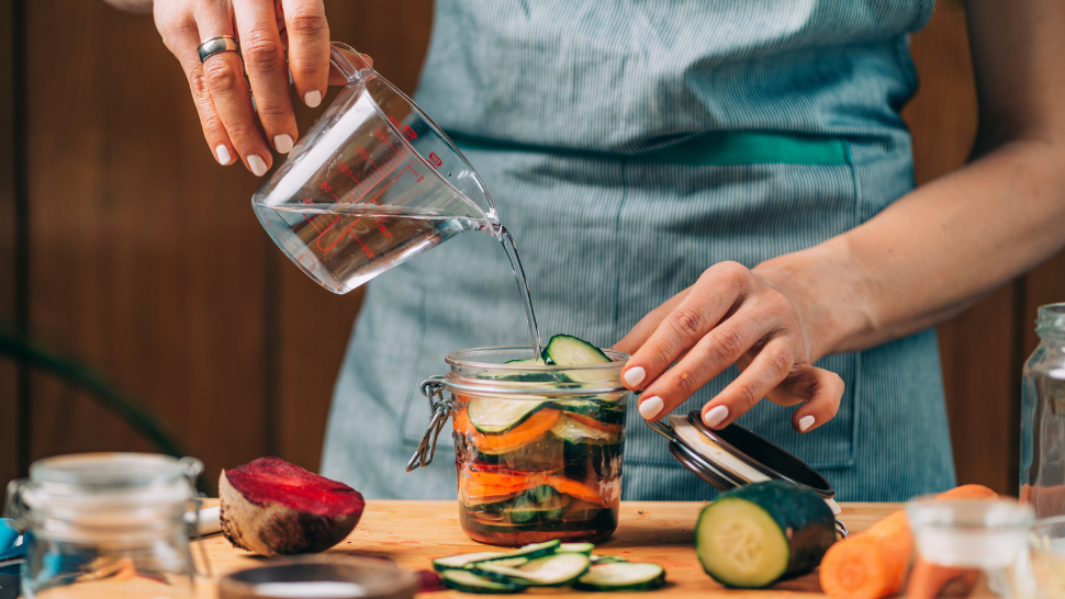 Person pouring water into a jar of sliced vegetables, preparing homemade pickled cucumbers and carrots.