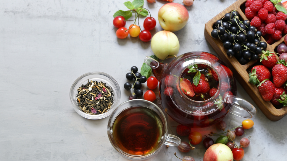 Glass teapot with berry tea surrounded by fresh fruit, loose tea leaves, and a cup of brewed tea.